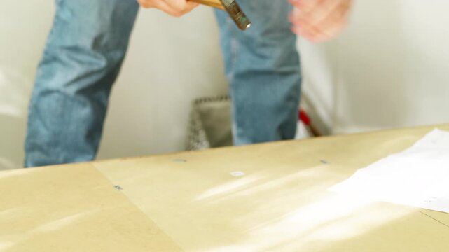 Male individual assembling furniture at home using a hammer to secure components on a wooden surface with assembly instructions visible in a bright indoor setting