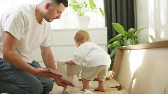 Father and toddler assembling furniture at home, with the child exploring cardboard pieces while the parent organizes materials in a bright living room setting