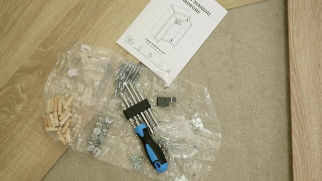 Tools and assembly components for furniture assembly at home, including screws, dowels, and a manual, arranged on a wooden floor with visible packaging