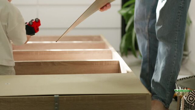 Adult assembling furniture at home with child nearby, placing wooden panels into a drawer structure in a bright, modern living space with plants and light colors