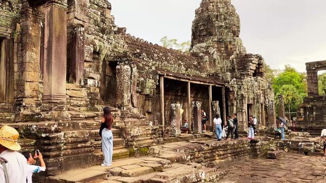 Scenic footage of visitors exploring the ancient Bayon Temple in Angkor Thom, Siem Reap, Cambodia. The video showcases the temple's iconic smiling stone face tower, weathered Khmer architecture, and