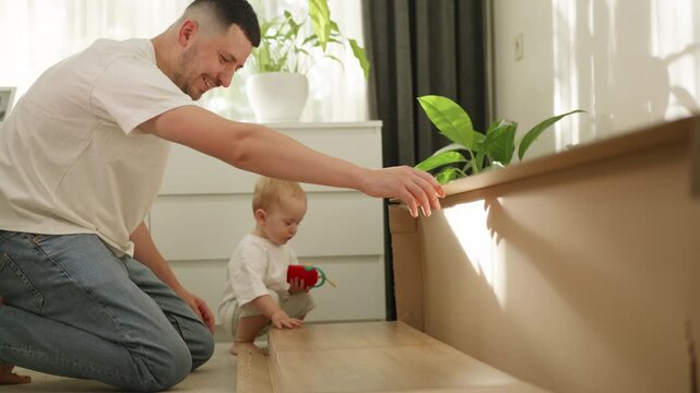 Father and toddler assembling furniture at home, with the child playing nearby, as the man positions wooden panels on a light-colored surface in a bright living room