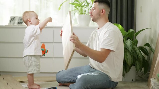 Father and toddler assembling furniture at home, with the child holding a toy while the parent secures a panel, surrounded by indoor plants and a bright living room setting
