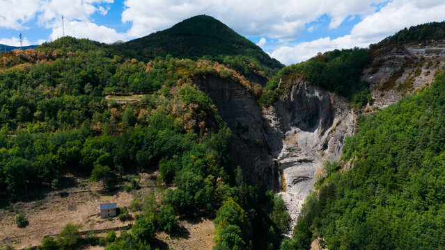 A winding hiking path traverses a vibrant, lush green mountain valley, framed by towering grey peaks and dense forest, under a bright blue sky 