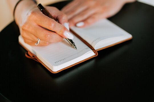 Woman writing in a small notebook with a black pen on a round wooden table during the daytime