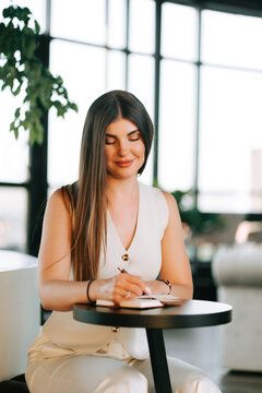 Young woman sitting at a round table and writing in a small notebook inside a bright modern office