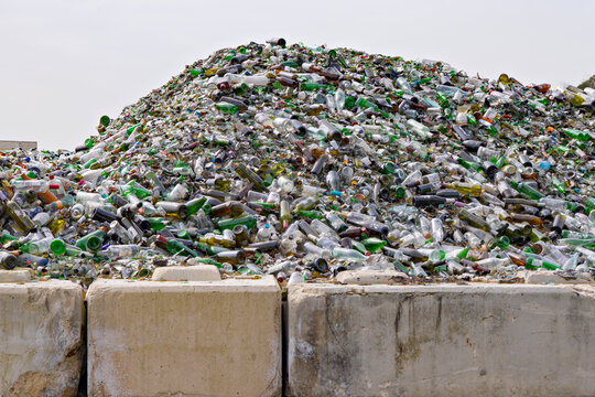 Huge pile of discarded glass bottles and broken containers behind concrete barriers at recycling facility, showing waste accumulation and material recovery