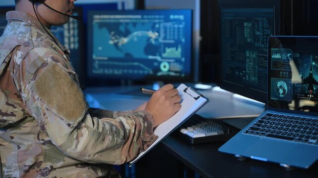 Military operator with headset analyzes radar HUD screens and takes notes in high tech command center