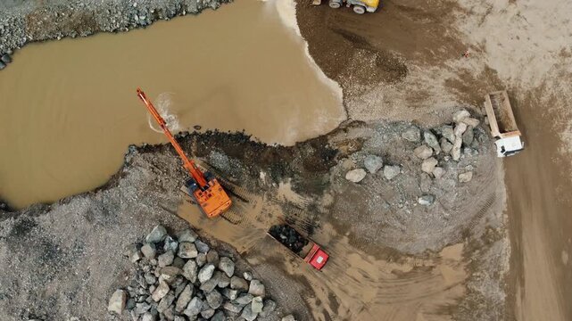Top-down aerial view of heavy machinery working on a riverbank construction site, with an excavator scooping rocks from the water and loading a dumper truck while a wheel loader moves earth
