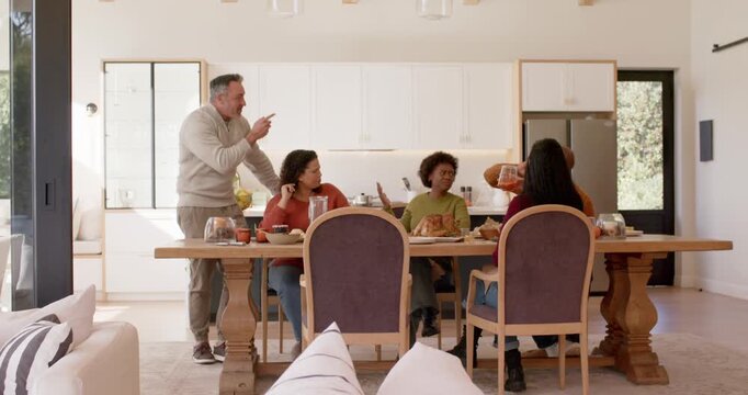 Diverse friends sitting at table in kitchen, standing man speaking, offering pitcher to share drink