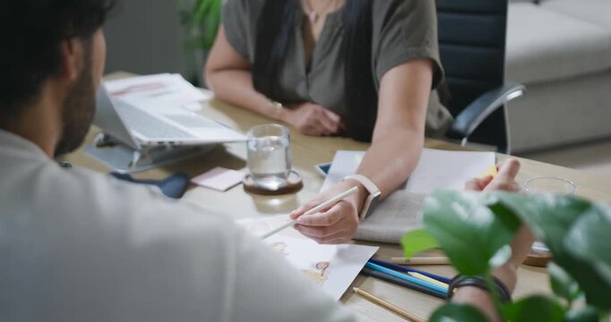 Woman is offering white pencil, man is reaching and marking sketches during design review at table