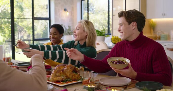 Three friends in sweaters passing roasted turkey and bowls at home table starting meal, laughing