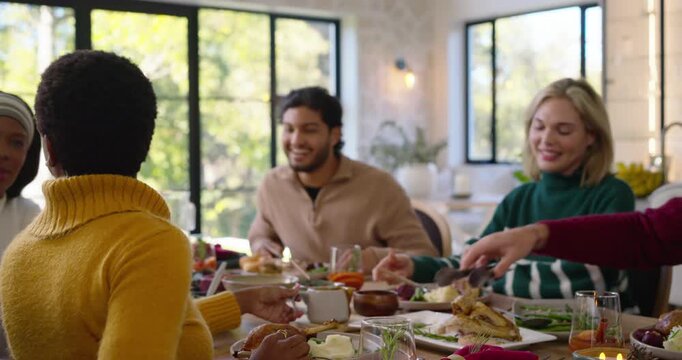 Diverse friends wearing sweaters sharing roasted bird at home table while green woman offering bite