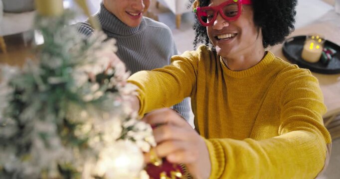 Two African American men decorating frosted holiday tree at home, grabbing ornaments from basket