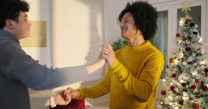 Diverse male couple on sofa by Christmas tree mustard sweater man sitting up clasping hands dancing