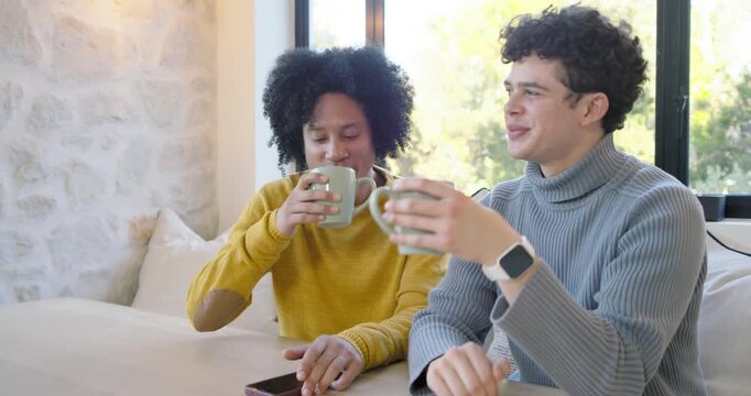 Diverse male friends sharing joke, smiling and sipping light-green mugs on couch by window