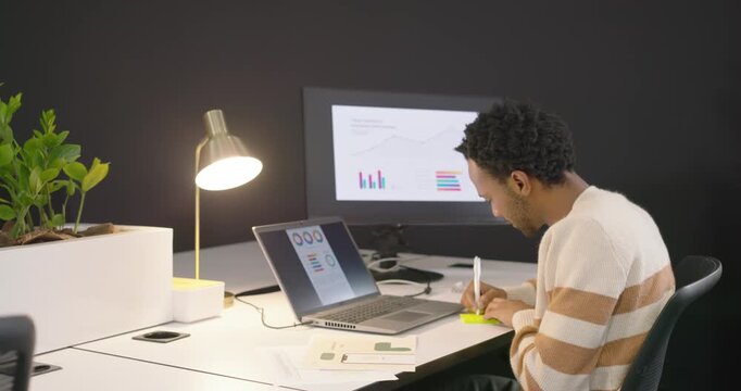 African American man glancing at laptop charts noting and comparing reports with monitor for review