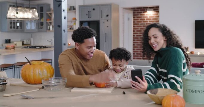 African American family gathering in kitchen woman showing phone man guiding child carving pumpkin