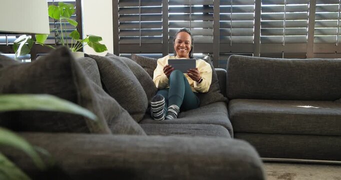 African American woman in yellow pullover reacting to tablet, smiling while holding it on gray sofa