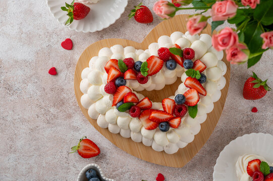 Valentine&rsquo;s Day heart-shaped Pavlova cake decorated with fresh strawberries, blueberries, raspberries and mint leaves on a wooden board. Romantic holiday dessert. Copy space.