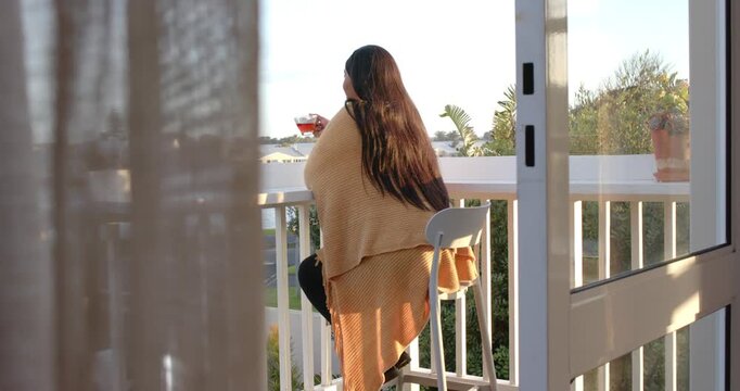 Curtain drifting in breeze nudging African American woman adjusting shawl on balcony, holding glass