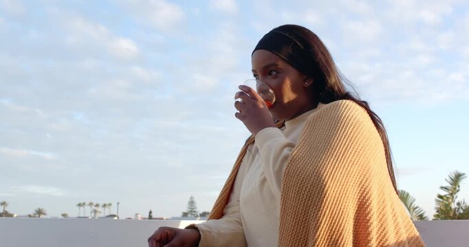 African American woman sipping cup, warming in mustard blanket gazing at skyline on roof