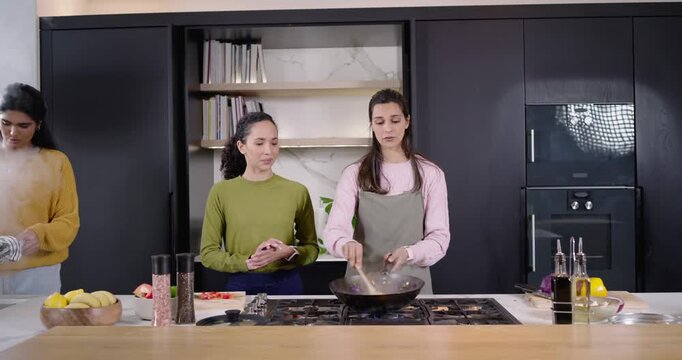 Diverse female cooks stirring wok at kitchen island, third bringing hot pot to set on stove