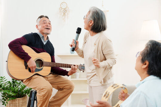 Cheerful, joyful Asian senior friends spending leisure time together. Elderly man playing acoustic guitar while senior women sitting on sofa singing along in white cozy, modern living room at home