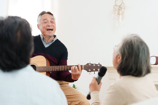 Cheerful, joyful Asian senior friends spending leisure time together. Elderly man playing acoustic guitar while senior women sitting on sofa singing along in white cozy, modern living room at home