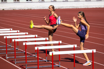 Fototapeta premium Adult and teenage female athletes lift legs over training hurdles. Concept of flexibility, dynamic stretching, coordination, high jump preparation and joint practice.