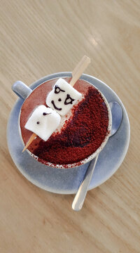 A rich and creamy hot chocolate served in a white ceramic mug, topped with fluffy marshmallows arranged to look like a playful cartoon face with a chocolate drizzle smile.