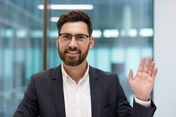 Close-up photo of a young man in glasses and a suit sitting in an office, talking and greeting the camera © Tetiana