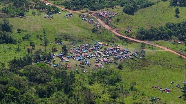 Drone footage soars above the lively WRC Rally 2026 campsite in Nueva Alborada, Paraguay, showing colorful tents, numerous cars, and spectators gathering in lush green fields under bright daylight.