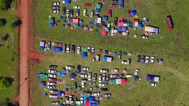 Spectators and campers gather beside a red dirt road near the Nueva Alborada rally stage in Nueva Alborada, Itapua, Paraguay, South America, with tents and parked vehicles, top-down drone forward fly