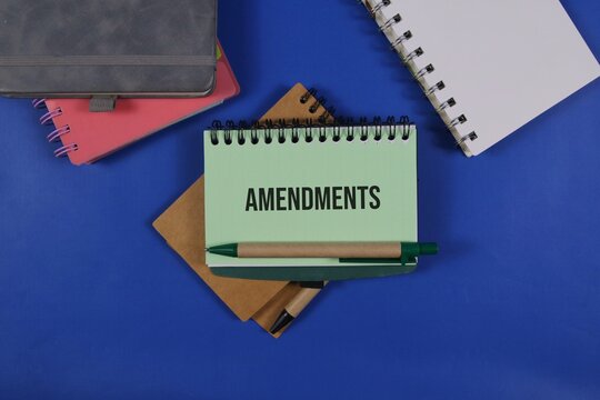 a spiral notebook with the word &ldquo;Amendments&rdquo; surrounded by colorful notebooks and a pen on an dark blue background, representing legal changes, document revision, policy updates, and business planning