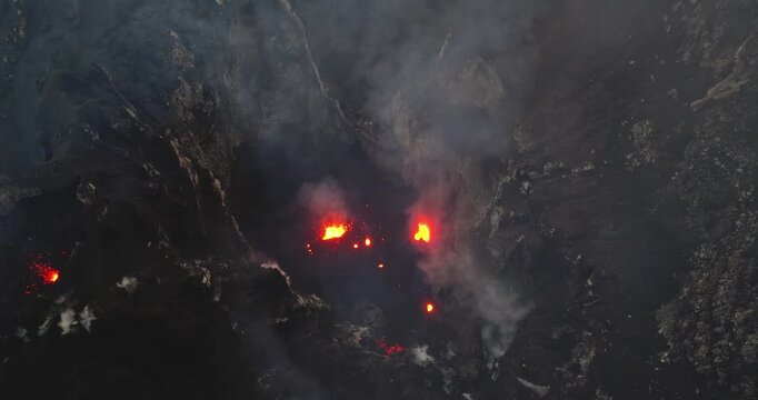 Aerial view discovering Mount Yasur volcano at night, crater actively erupting, spewing glowing lava, magma, and dense smoke into a dark, rocky landscape on Tanna Island, Vanuatu. Drone panorama shot