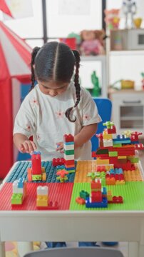 Girl with braided hair leaning over table, hands stacking multicolored plastic building blocks and small bricks on playboard in building; concentration creativity.