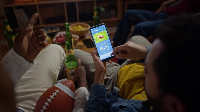 Multiethnic young adult male football fans placing online bets on smartphone while watching game at home. Ideal for mobile sports betting commercials and digital gambling marketing