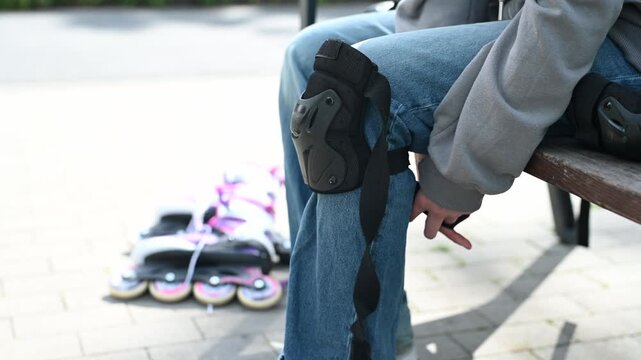 Skater wears knee pads, sits on park bench.