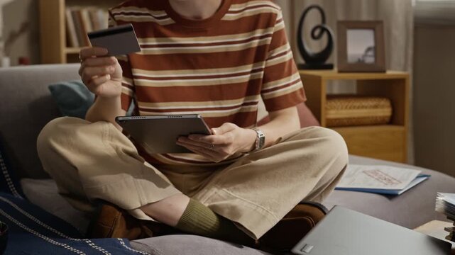 Young adult Caucasian woman sitting on couch, holding credit card and typing on tablet to pay bills, camera tilting up. Clip is ideal for online banking and digital payment advertisements