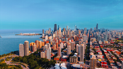 Obraz premium Chicago cityscape along Lake Michigan shoreline. Aerial photograph of dense city buildings, green parkland, blue coastal water and clear horizon under bright light.