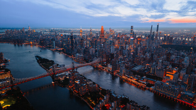 Twilight view of Midtown Manhattan skyline and Queensboro Bridge on East River. Illuminated skyscrapers and bridge reflect orange lights into dark river water.