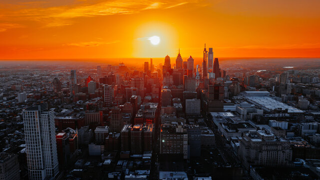 Skyline silhouette of Philadelphia during bright orange sunset. Sun sets directly behind tall office towers casting warm glow over urban landscape.