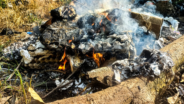 An eye-level photograph capturing a pile of trash and household waste&mdash;including cardboard, paper, and egg cartons&mdash;actively burning on the ground.