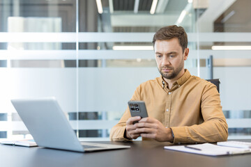 Fototapeta premium Businessman concentrating on his smartphone while sitting at a sleek desk with a laptop and documents, engaging with digital communication and technology in a corporate environment