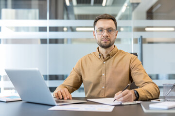 Obraz premium Confident businessman working diligently at a modern office desk, wearing glasses and a casual shirt, while typing on a laptop and taking important notes on paper