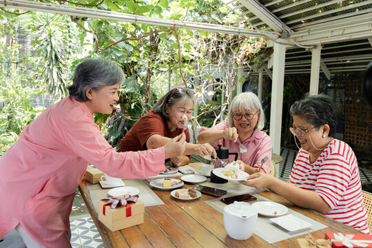 Retired friends keep sharing bites of cake at a garden cafe, laughing over the plate during a lighthearted reunion that highlights affection, conversation and pleasure in togetherness, aging society.