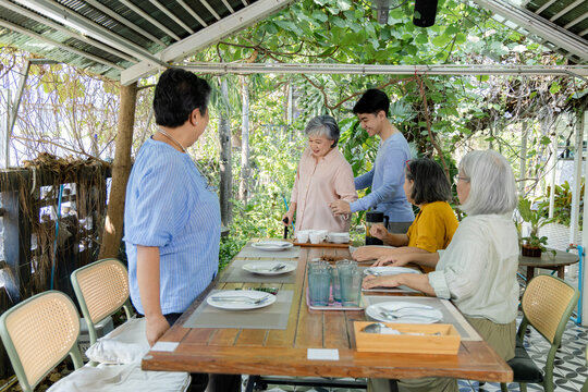 An elderly woman arrives at an outdoor cafe with support from a young man while friends wait at the table, showing caring assistance, mobility support and a warm reunion spirit today, aging society.