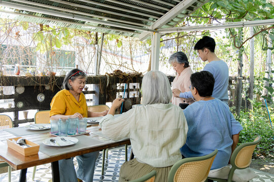 A waiter stands by the outdoor cafe table with a tablet, taking orders from senior friends during a reunion where conversation, service and mealtime choices bring everyone together, aging society.