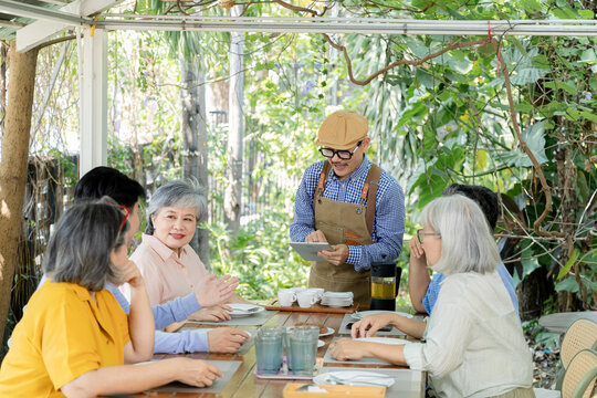 A smiling waiter confirms an order on a tablet while senior friends chat around an outdoor cafe table, creating a cheerful scene of service, companionship and relaxed reunion dining, aging society.